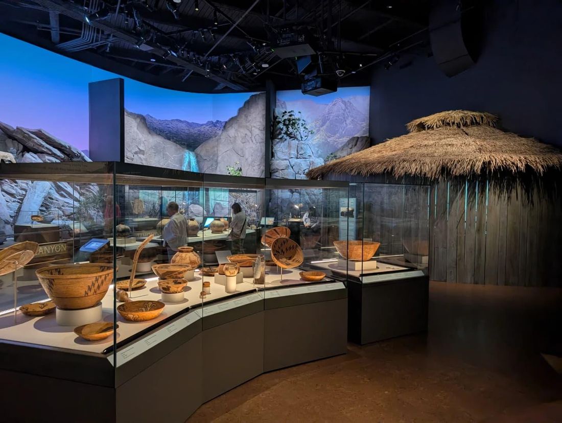 A dim museum gallery: glass cases of woven baskets and clay pots curve beneath a projected mural of a desert canyon and waterfall, beside a round shelter with a thatched roof.