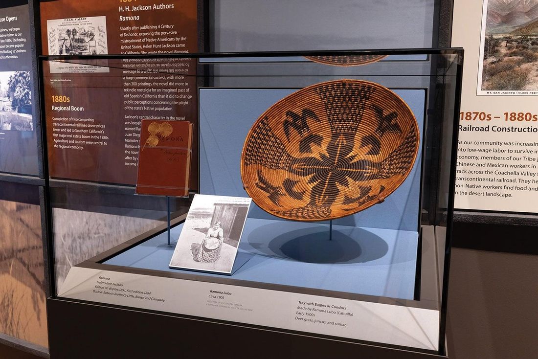In a glass case, a coiled basket tray patterned with dark birds stands on a mount beside a black-and-white photograph of a seated woman with a basket, exhibition panels behind.