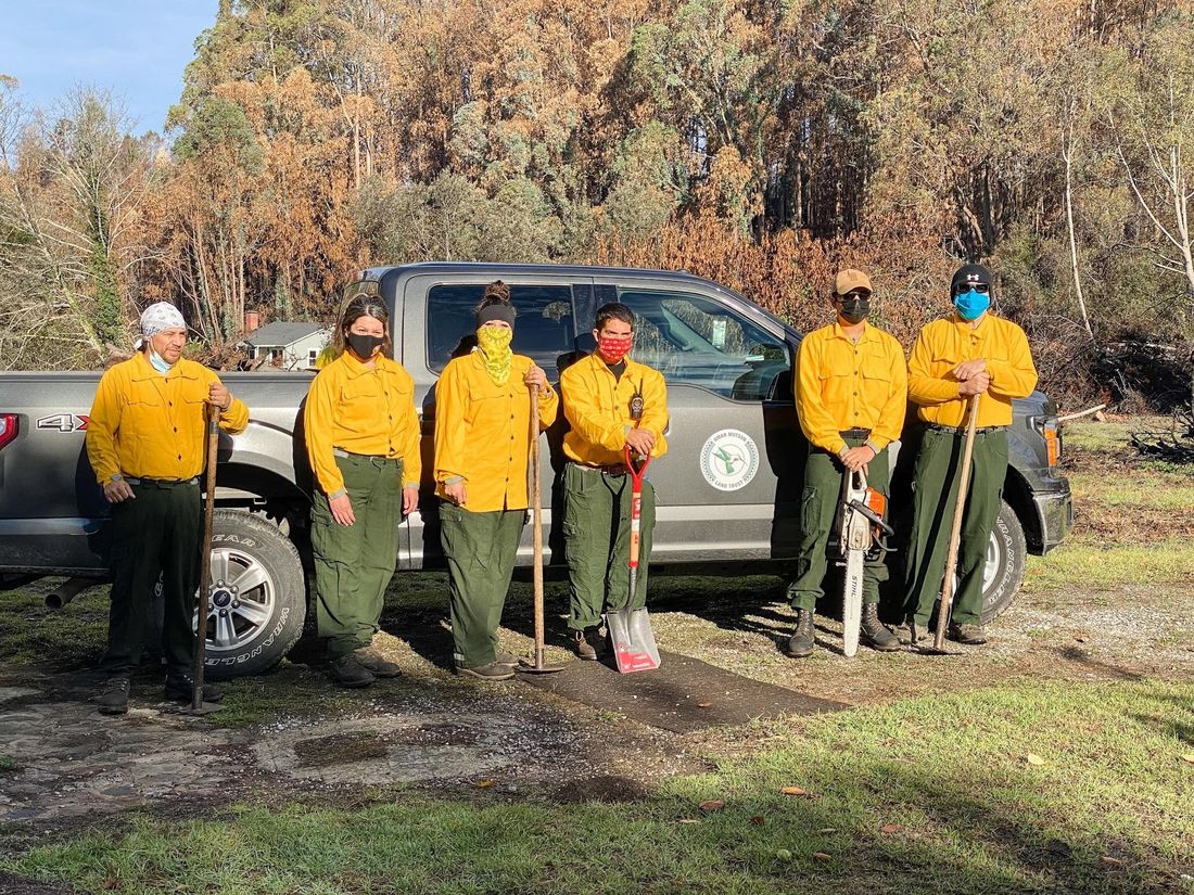Six people in yellow fire shirts and green trousers stand with hand tools in front of a pickup truck, fire-scarred trees behind them.