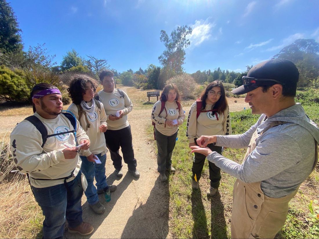 Rick Flores, in a cap, holds out a plant cutting to a group of young people in matching sweatshirts on a dirt path through coastal scrub.