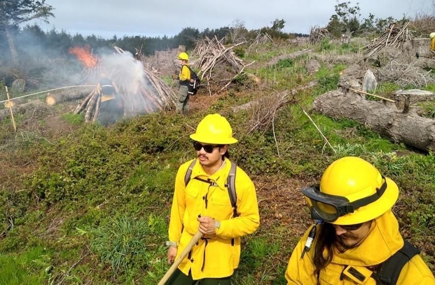 Two people in yellow fire gear and hard hats stand on a green slope in the foreground while a pile of brush burns and smokes behind them.
