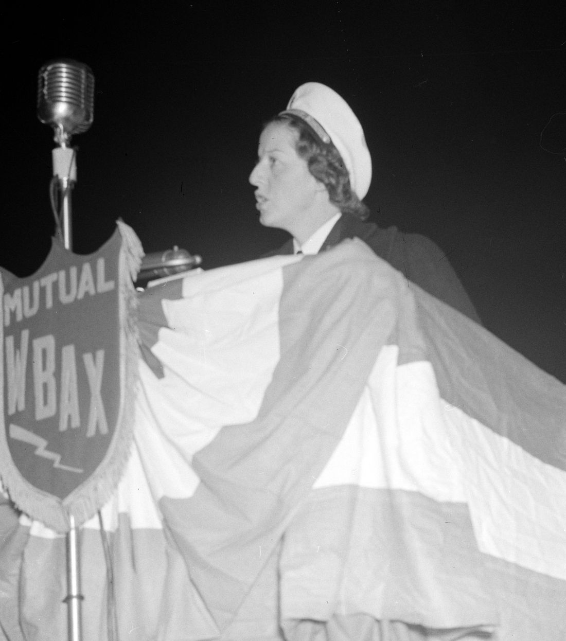 Ann Bernatitus in a white Navy nurse's cap speaks at a microphone on a stage draped with bunting, beside a radio station's shield reading Mutual WBAX.