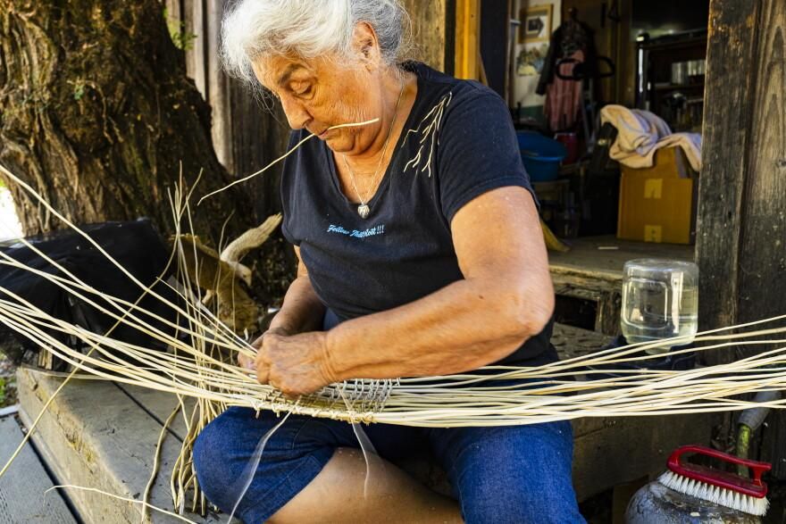 Wilverna Reece, a woman with white hair, sits on a porch weaving a basket, long pale strands of fiber fanning across her lap.