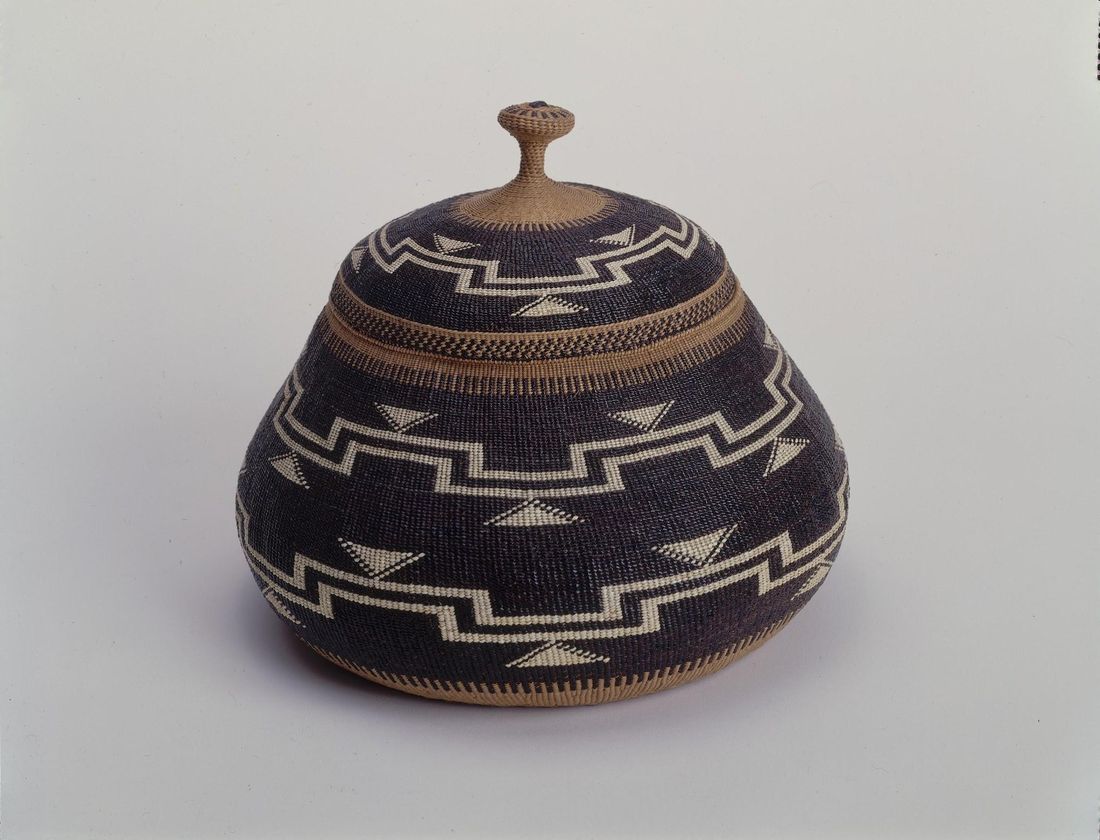A rounded lidded basket woven in dark purple-brown with bands of white zigzags and triangles and a small knob on its lid, photographed on a plain white ground.