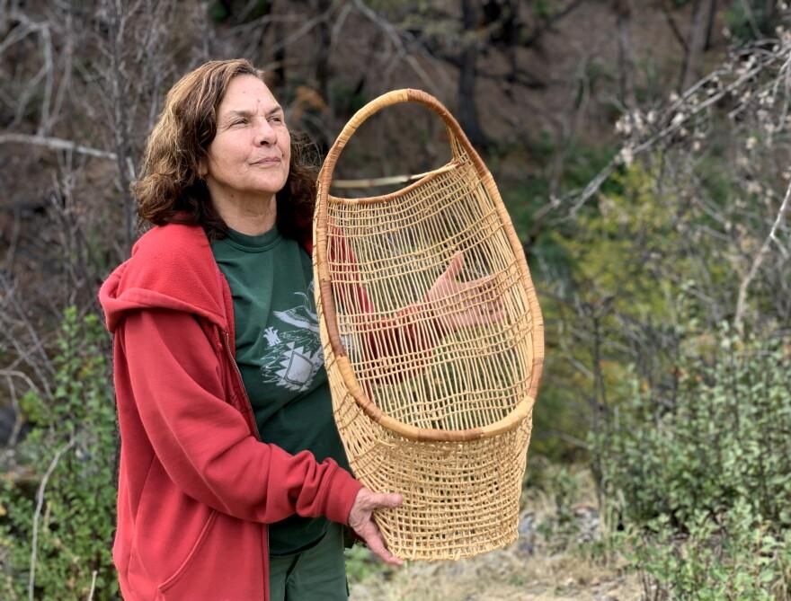 Kathy McCovey, in a red hooded sweatshirt, holds up a tall open-weave basket outdoors, bare branches and green shrubs behind her.