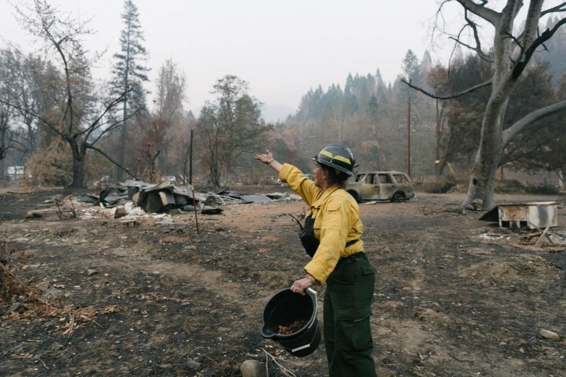 Kathy McCovey, in a yellow fire shirt and helmet, holding a bucket, throws acorns across a burned clearing with charred trees, a burned-out car and the remains of a building.