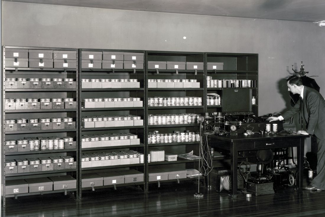 An attic room lined with shelves of boxed wax cylinder recordings; a person in a dark suit bends over a table holding a phonograph and recording equipment.