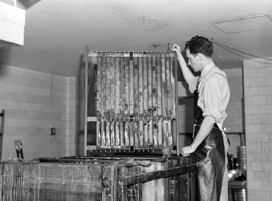 A man in a work apron lifts a rack of developed microfilm out of a processing tank in a tiled laboratory.