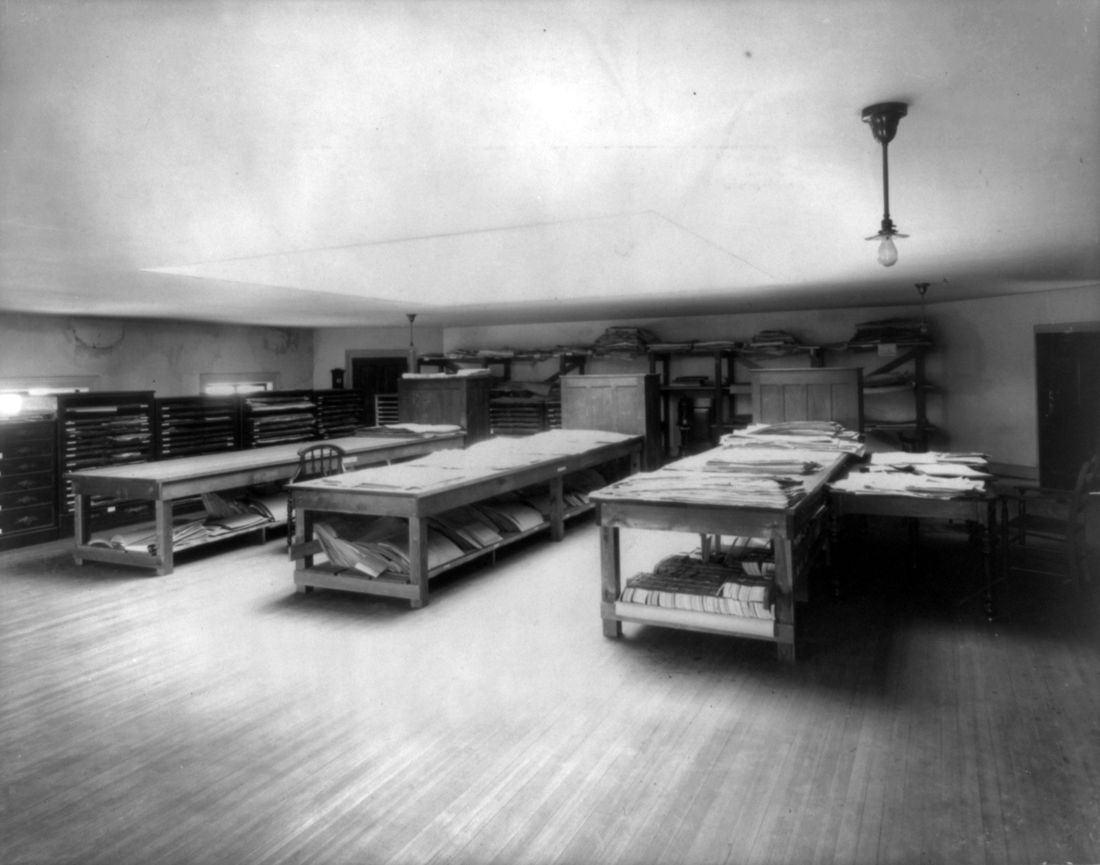 An attic room with long wooden tables and shelves stacked with prints and portfolios, flat-file cabinets along the wall and a single hanging lamp.