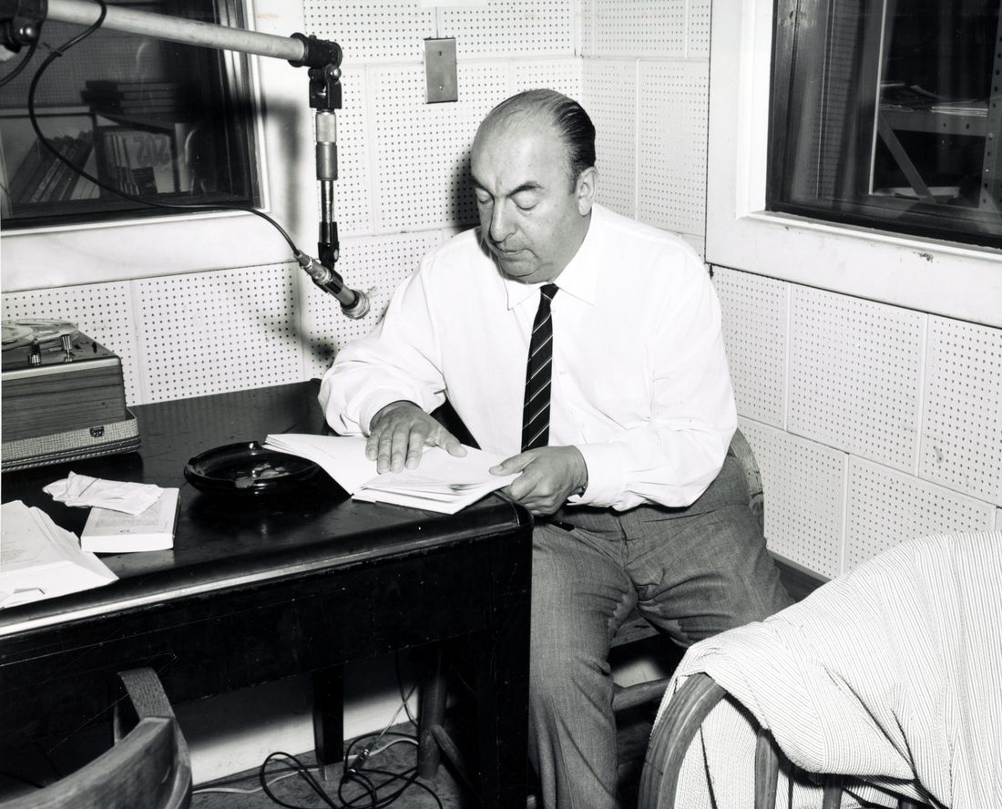 A man in a white shirt and tie reads from papers at a desk in a recording studio, a microphone on a boom above him and a disc on the desk.