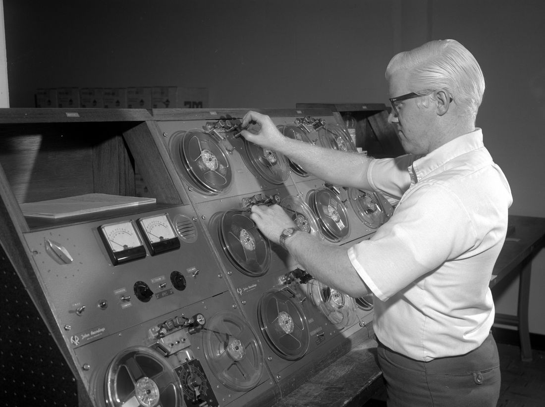 A man threads magnetic tape on a console of reel-to-reel recorders, copying a master tape.
