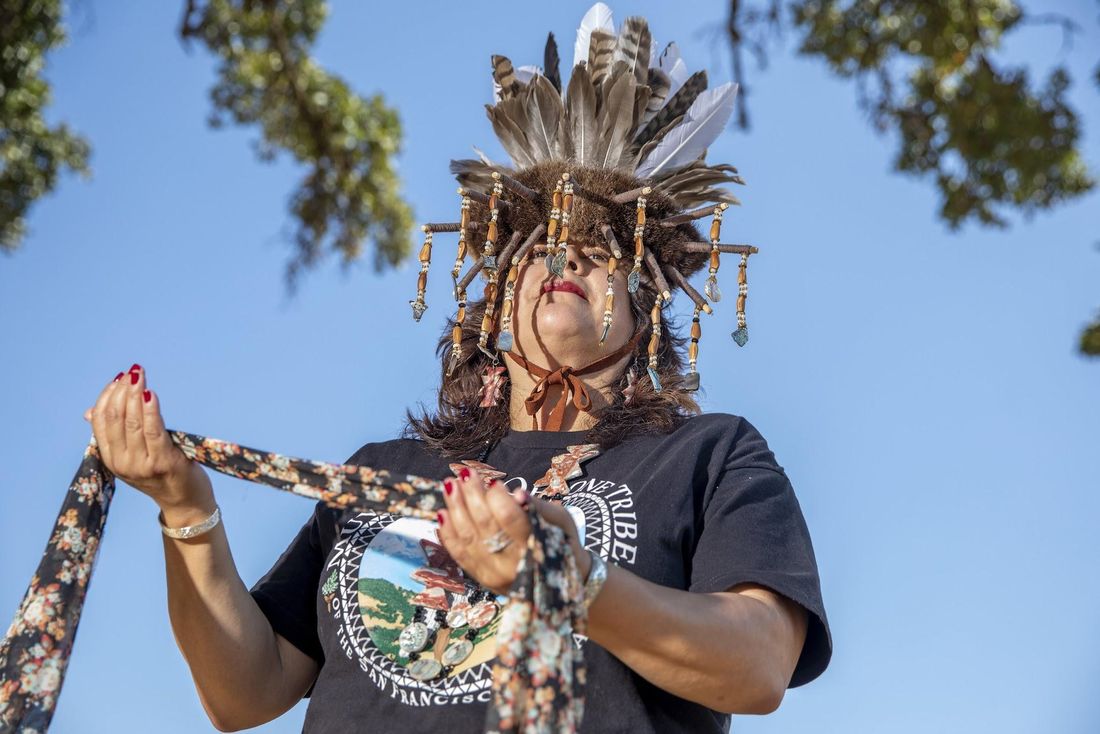 Seen from below against a blue sky, Monica V. Arellano, in a feathered headdress hung with beaded strands, holds a floral scarf, wearing a black tribal T-shirt.