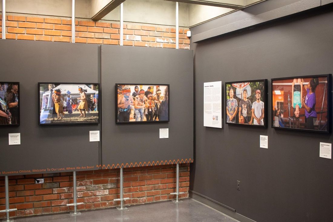 Framed color photographs of community members hang on dark gray panels in a gallery with brick walls and a concrete ceiling.