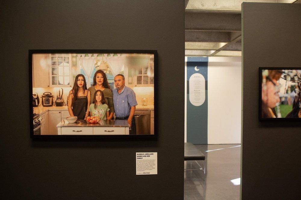 A framed color photograph of a family of four standing in their kitchen hangs on a dark gallery wall beside a small printed label.
