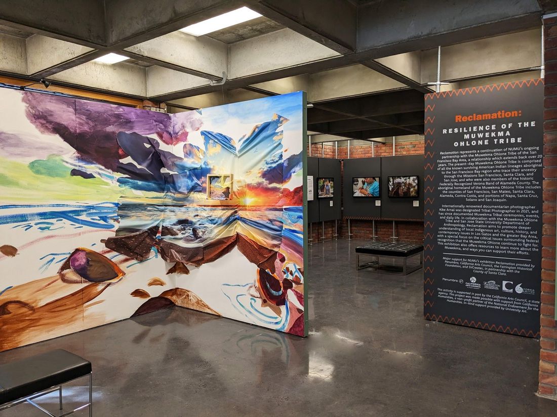 A tall painted panel of swirling color stands at the entrance to a gallery, beside a dark title panel and walls of framed photographs.