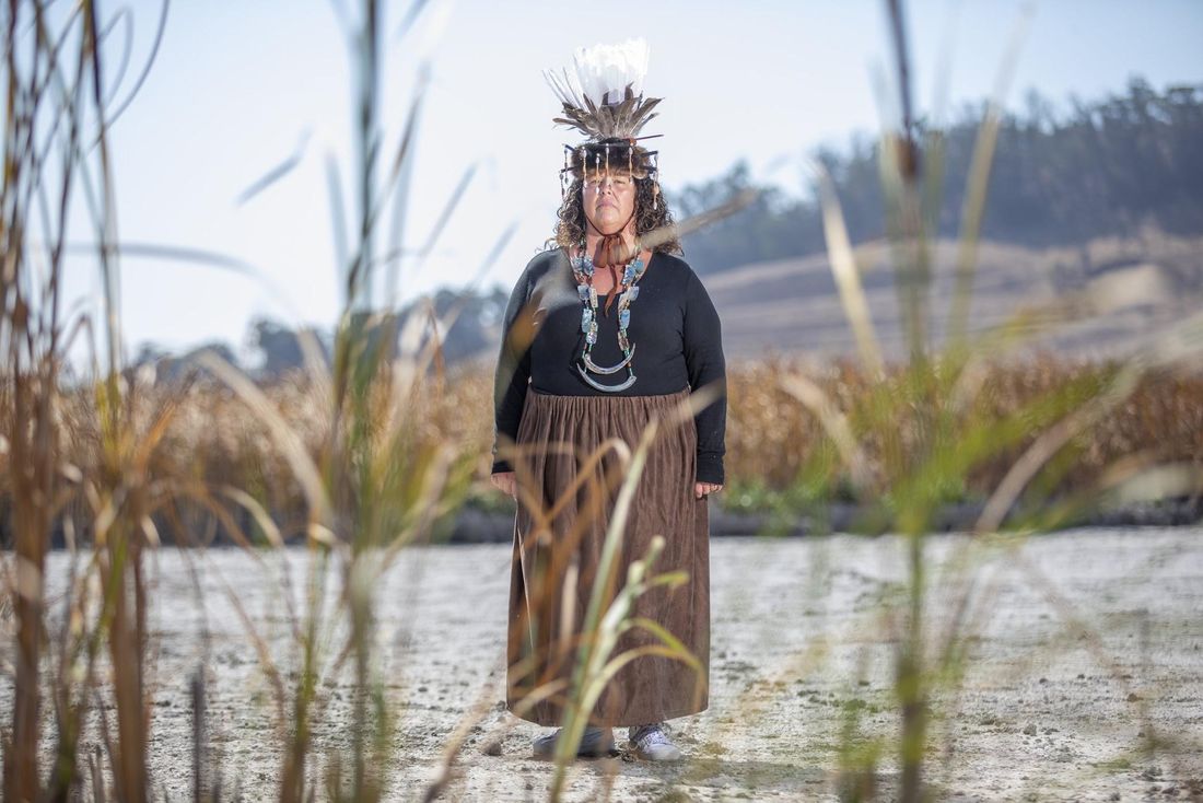 Sheila Guzman Schmidt, in a feathered headdress, shell necklaces, a black top and a long brown skirt, stands on dry pale ground framed by tall grasses, hills behind her.