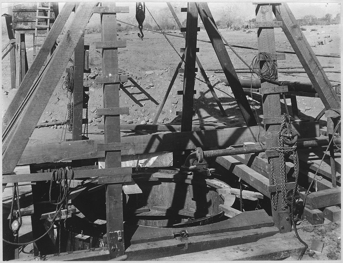A timber headframe with pulleys, chains and rope over a round well casing under construction, on a flat of river cobbles with brush and low hills behind.