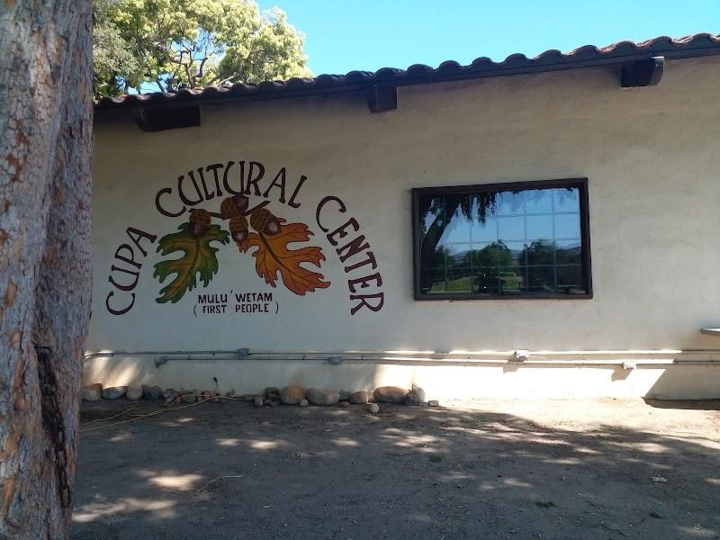 A white adobe wall painted with the words Cupa Cultural Center arched over three oak leaves, and Mulu' Wetam (First People) beneath, beside a window in the shade of a tree.