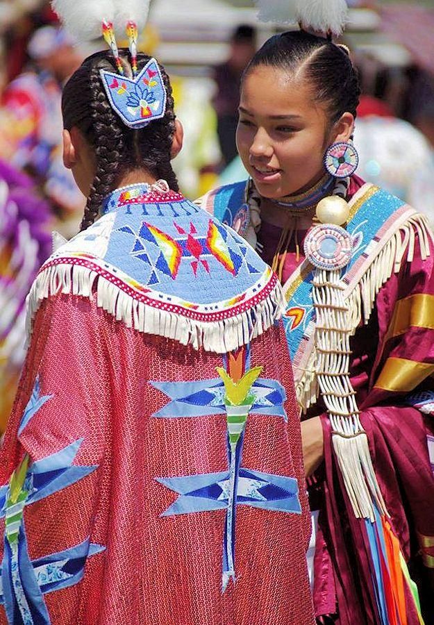 Two young women in beaded regalia at a powwow: one, seen from behind, wears a red cape with a beaded collar and long fringe; the other looks aside, smiling, in large beaded earrings.