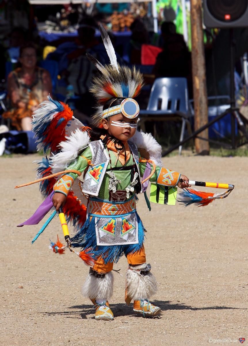 A small child in dance regalia, with a feathered headdress, beaded vest and feather bustle, dances across a dirt arena holding a decorated staff, spectators under shade behind.