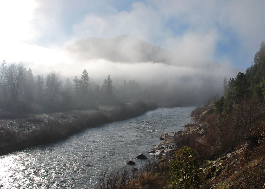 Morning fog lifting off a forested ridge above the Klamath River, which runs over a wide gravel bar between bare winter trees and a steep, pine-covered bank.