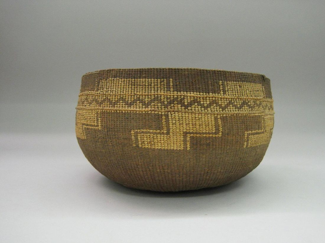 A deep round basket woven in tan and gold, with a stepped geometric band and a zigzag line below the rim, on a pale gray ground.