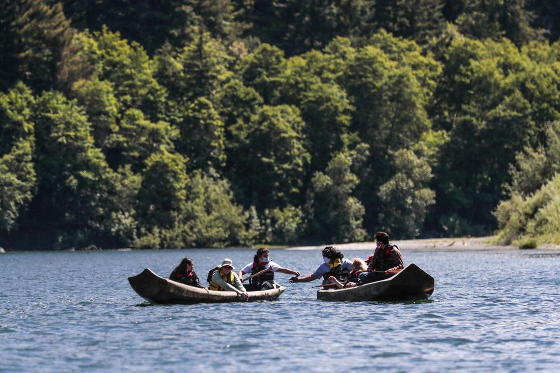 Two dugout canoes with paddlers in life jackets meet on a blue river beneath a steep, densely forested bank.