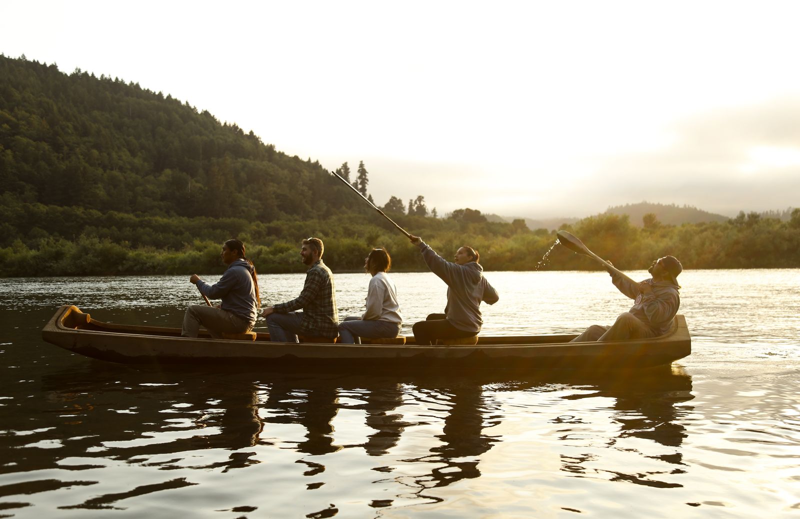 Five people paddle and pole a long wooden canoe across the calm Klamath River at sunset, forested hills behind them and golden light on the water.