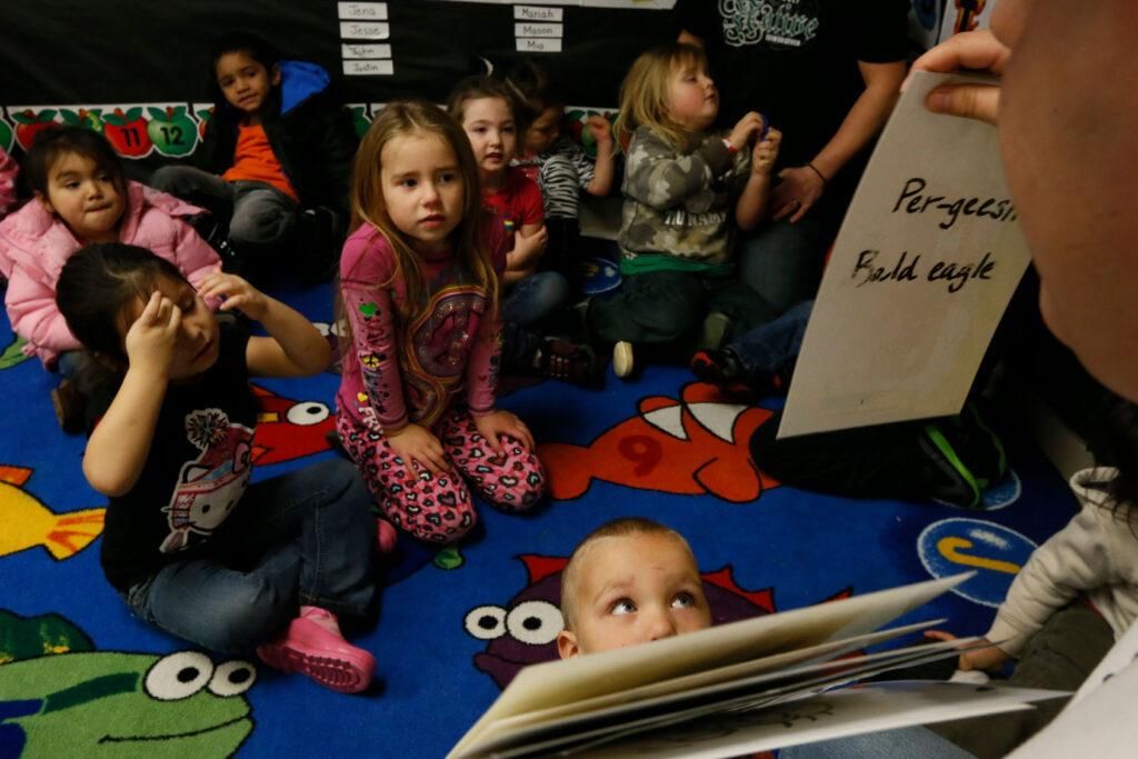Preschool children sit on a bright classroom rug and look up at their teacher, who holds a handwritten card with a Yurok word above the words Bald eagle.